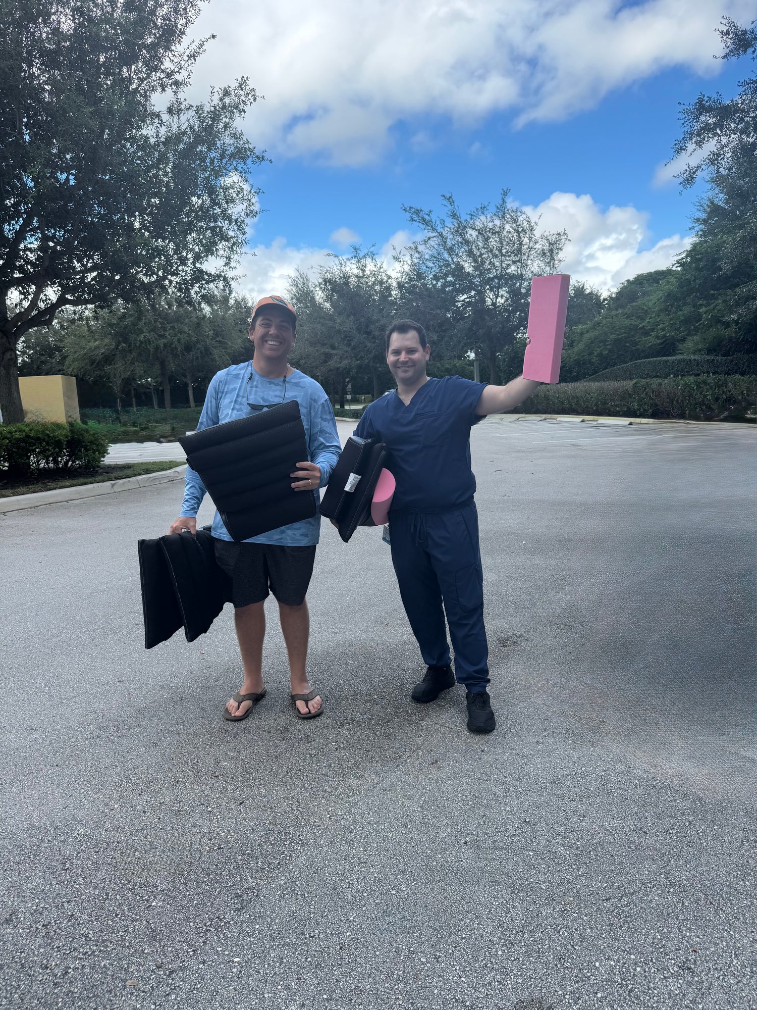 Two people, Spencer and Jake, the inventors of the Backliner are happily holding Backliners and pink foam which is a key component of the Backliner in a parking lot with trees and blue sky in the background.